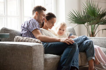 Young family with daughter using app or watching educational video holding phone sitting on couch at home, married couple with little girl focused on smartphone doing mobile shopping or checking news