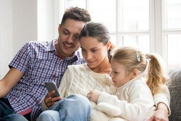 Obraz premium Parents with daughter watching video on mobile phone at home, young family couple with little girl using app for education or checking social networks focused on smartphone sitting together on sofa