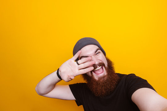 Cheerful young bearded man holding hand on face and taking selfie