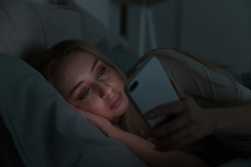 Portrait of young sleepy tired woman lying in bed under the blanket using smartphone at late night,...