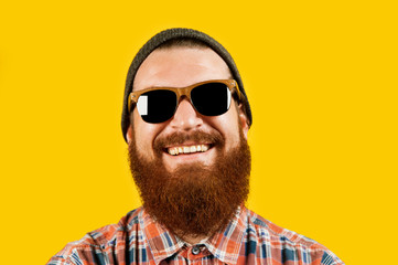 Obraz premium Portrait of young hipster man in sunglasses and hat posing on yellow background. Smiling bearded man wearing sunglasses and looking confident at the camera, studio shot.