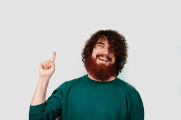 Smiling bearded hipster man with curly hair pointing away with bright smile on white background.