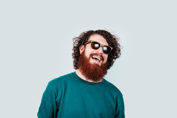Portrait of cheerful bearded hipster man with long curly hair waring sunglasses and smiling over white background