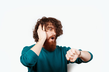 Portrait of a shocked bearded hipster man with long curly hair looking at his watch isolated over white background