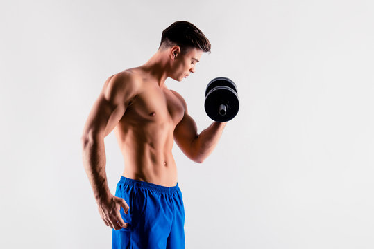 Side profile view photo of handsome sportive strong-willed naked muscular guy building muscles on arms by lifting dumbbells, clothed in blue sportive shorts, isolated on grey background
