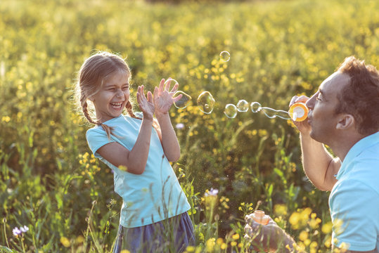 Joyful father is blowing soap bubbles in front of his laughing daughter. They are playing on summer meadow