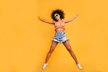 Hooray! Free hugs! I'm full of bliss! Attractive cheerful joyful careless mulato fumping up woman with funny curly hairdo wearing denim skirt and knitted beige top isolated on yellow background - obrazy, fototapety, plakaty