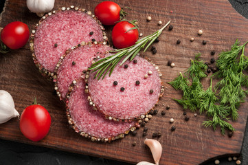Smoked salami on a black table and a brown board. Sausages with rosemary, garlic and pepper and tomato cherry, dill, parsley.