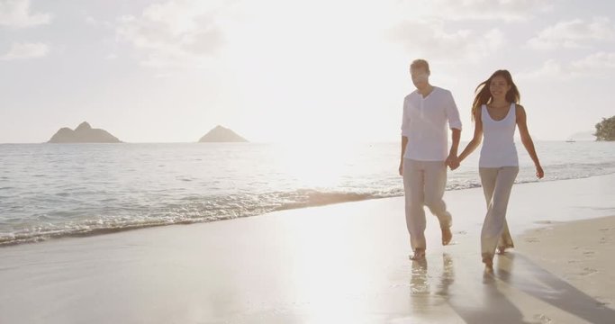 Beach sunset couple walking romantic on honeymoon on Hawaii. Sunrise romance young couple in elegant casual clothing walking together enjoying travel vacation holidays on Lanikai beach, Oahu, USA.