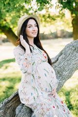 portrait of a pregnant girl in an embroidered beige dress and in a straw hat sits on a curved tree and wounds her hair