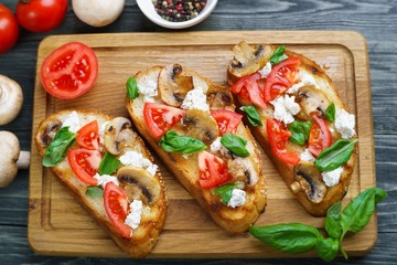 Tasty homemade Italian antipasti bruschetta with chopped tomatoes and champignons, garnished with ricotta and basil, healthy and delicious snack top view on wooden table