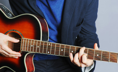 Fototapeta na wymiar Young hipster moustached man in casual jacket sitting on chair playing guitar on blue background. Practicing in playing guitar concept. Handsome young men playing acoustic guitar. Close up view.