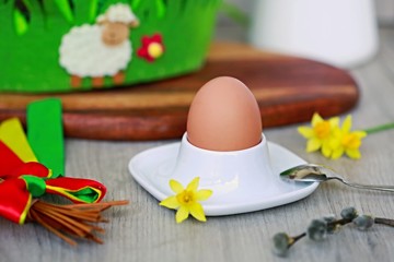 Colorful Easter decoration with hard boiled brown egg in a bowl, wooden table, spring yellow flower of daffodil, pussy willow twig, white lamb, red, green and yellow ribbon, white jar with milk