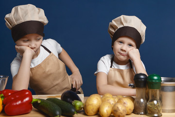 Portrait of two little boys dressed in chef uniform having bored looks, leaning on their elbows, don't want to help mother cooking dinner, standing at kitchen table with fresh organic vegetables