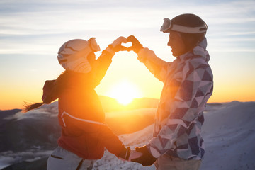 Lovely couple holding hands in shape of heart on snowy peak at sunset. Winter vacation