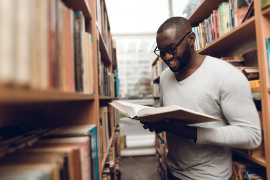Ethnic african american guy reading book, smiling in aisle of library.