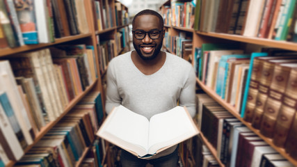 Ethnic african american guy reading book, smiling in aisle of library.