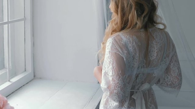 Young slim sexy blond girl in lingerie and boudoir standing near windowsill and looking in window.
