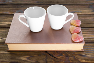 two cups on a book with jelly candies in the form of a heart on a wooden background.