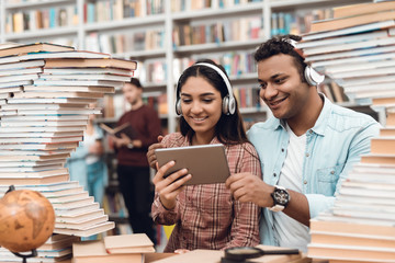 Ethnic indian mixed race girl and guy surrounded by books in library. Students are using tablet.