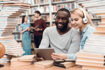 Fototapeta premium Ethnic african american guy and white girl surrounded by books in library. Students are using tablet.