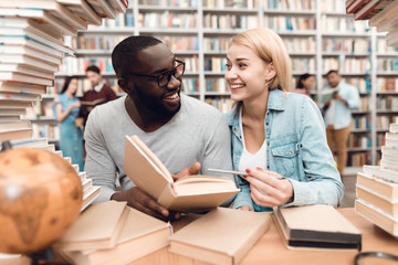 Ethnic african american guy and white girl surrounded by books in library. Students are reading book.