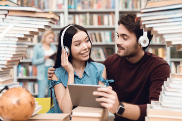 Ethnic asian girl and white guy surrounded by books in library. Students are using tablet with headphones.