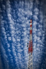 vertical image of a red and white communications tower with long clouds and sky in the background