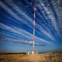square image of a red and white communications tower with long clouds and sky in the background