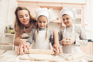 Children with mother in kitchen. Mother is helping kids to roll out dough.