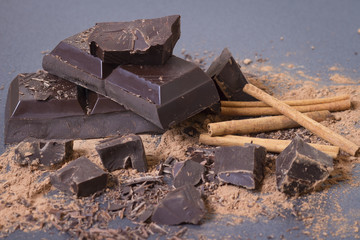 Broken pieces of dark chocolate close-up. Sweet chocolate background. Cocoa powder and cinnamon sticks on a black background