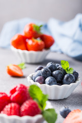 Fresh ripe raspberries, strawberries and bluebetties in white bowls on light gray concrete background. Healthy food concept.