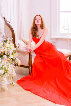 Magnificent young woman in luxurious red dress and precious jewelery posing in a luxury apartment. Classic vintage interior. Beauty luxury fashion 