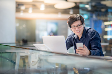 Young Asian business man listening to music by headphones and smartphone while working with laptop computer in co working space. freelance or digital nomad lifestyle concepts