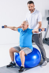 Obraz premium rehabilitation therapist assisting senior man exercising with dumbbells on fitness ball
