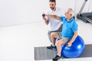Obraz premium high angle view of rehabilitation therapist assisting senior man exercising with dumbbells on fitness ball
