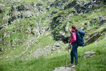 Young woman backpacker with trekking sticks and a backpack is standing on a stone at the foot of the Fagaras Mountains, Romania. Girl with trekking stick shows to the distance.