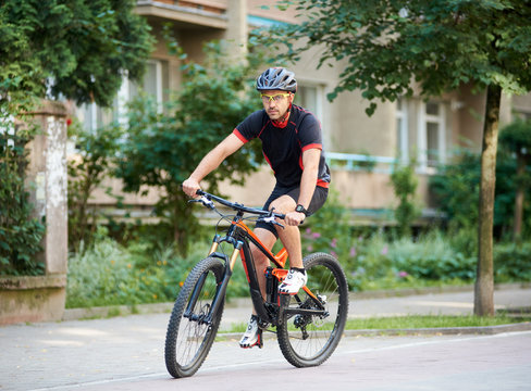 Sportsman in professional cycling garment and helmet looking to camera, riding bicycle near beautiful buildings. Man training, improving hobby, getting ready for contest