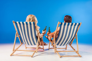 Rear view of young couple with beer bottles relaxing in deck chairs on blue background