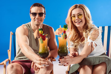 Young smiling couple with cocktails resting in deck chairs on blue background