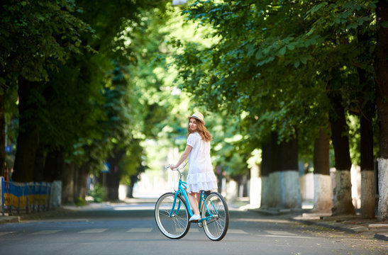 Green urban plantings. Female rides on retro bike alone at road on a summer day. The girl is dressed in a light dress and hat on her head