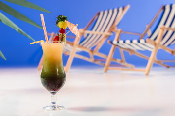 Glass with tropical cocktail by deck chairs on blue background