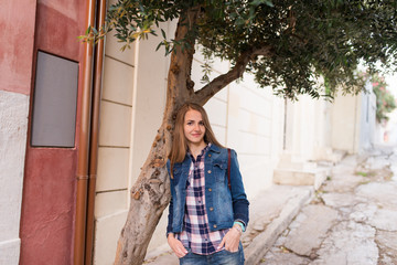 Smiling Caucasian girl. A girl with long hair in a denim suit leaned her elbows on the olive tree. Summertime