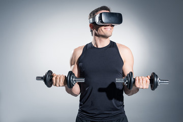 Obraz na płótnie Canvas Active man training with dumbbells while wearing vr glasses on grey background