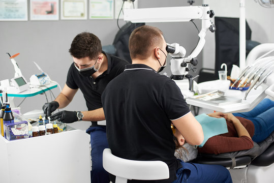 One male dentist is fixing a patient's teeth while another male dentist is preparing the dental filling