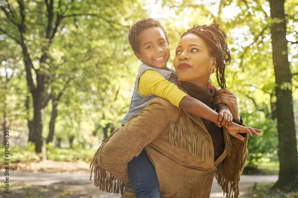 Wall mural  African American mother carrying his daughter on piggyback in park.