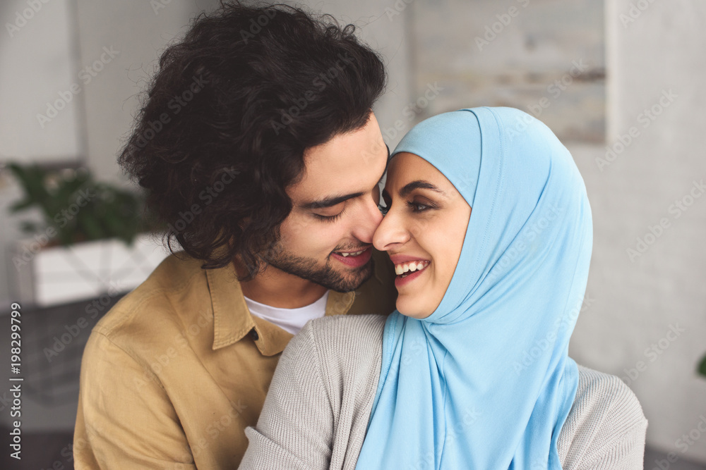 Wall mural smiling muslim couple hugging and touching with noses at home