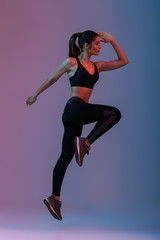 Fototapeta na wymiar Full length portrait in profile of female fitness instructor wearing black tracksuit doing aerobics in gym, isolated over purple background