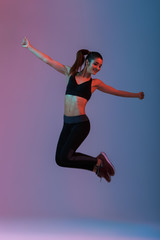 Fototapeta na wymiar Full length portrait of happy woman wearing black tracksuit jumping with hands aside during aerobics in gym, isolated over purple background