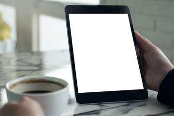 Mockup image of hands holding black tablet pc with white blank screen and coffee cup on table background